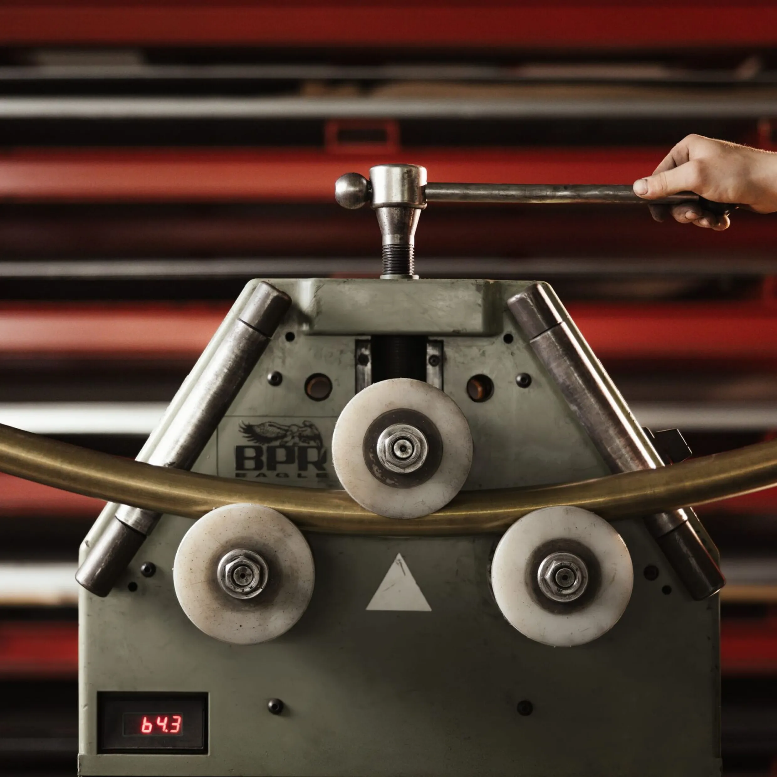 Rohrbiegemaschine in einer Metallwerkstatt; eine Person bedient den Hebel, ein gebogenes Rohr führt durch Rollen, rote Regale im Hintergrund.