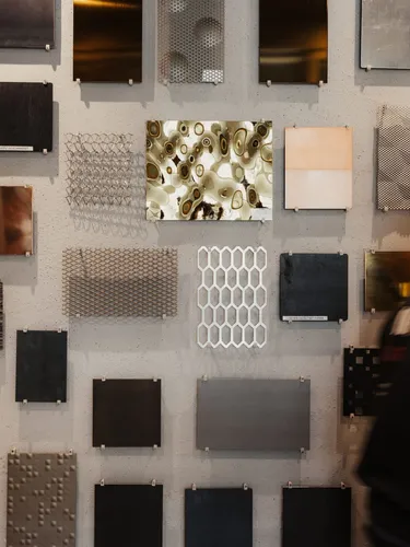 Wand mit zahlreichen Metallproben und Oberflächenmustern in quadratischer Anordnung; eine unscharf vorbeigehende Person in dunkler Kleidung und Mütze.