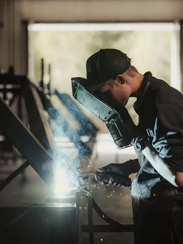 Person beim Schweißen in einer Südtiroler Metallbau-Werkstatt, Funken fliegen, Schutzhelm und Handschuhe, arbeitet an einer Metallkonstruktion.