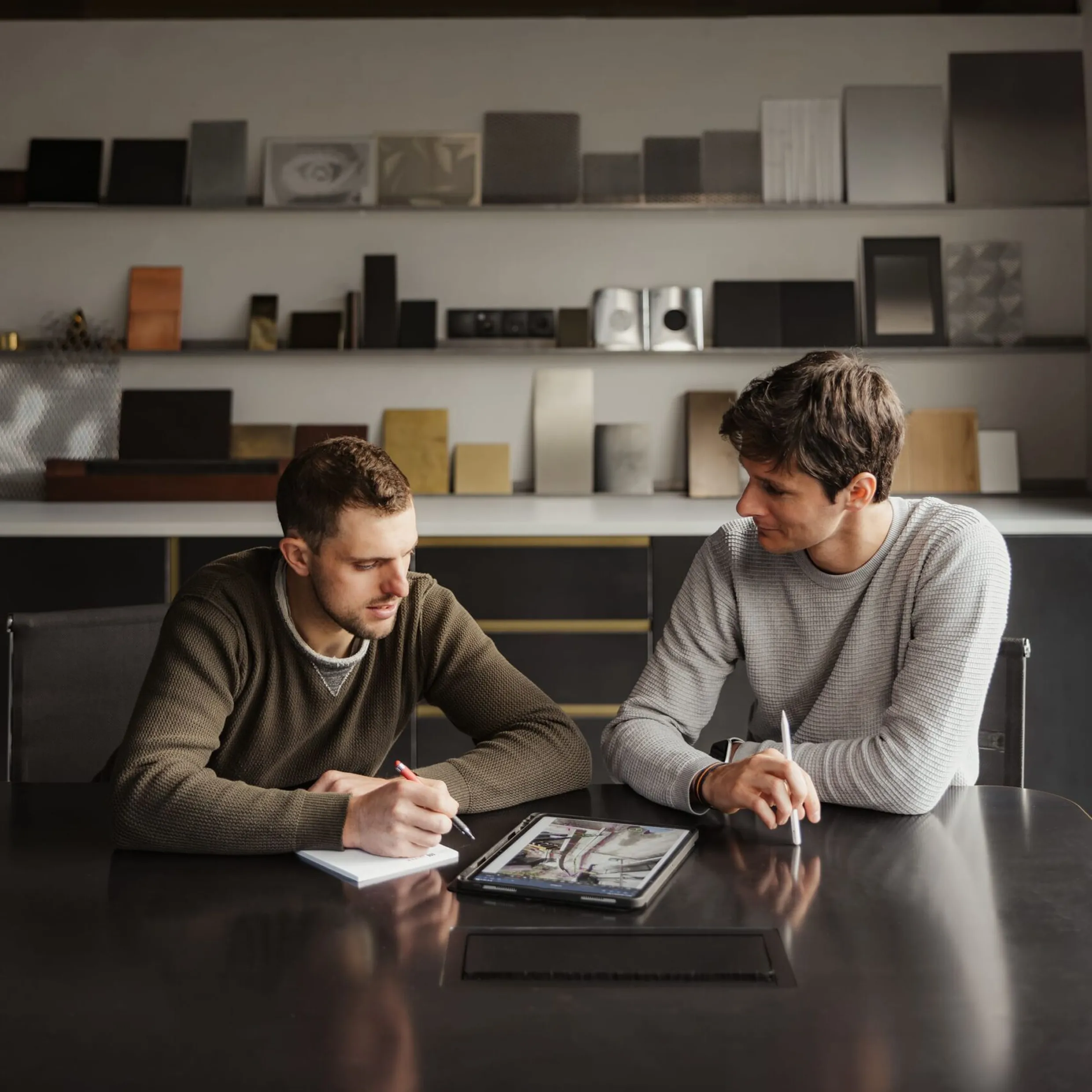 Zwei Personen aus Südtirol arbeiten an einem dunklen Tisch, notieren, schauen auf ein Tablet mit Entwürfen, Metallproben stehen auf Regalen.