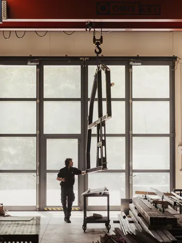 Metallbau-Werkstatt in Südtirol: Eine Person in dunkler Arbeitskleidung bedient einen hängenden Metallrahmen vor großen Glastüren; rechts Regale mit Profilen.