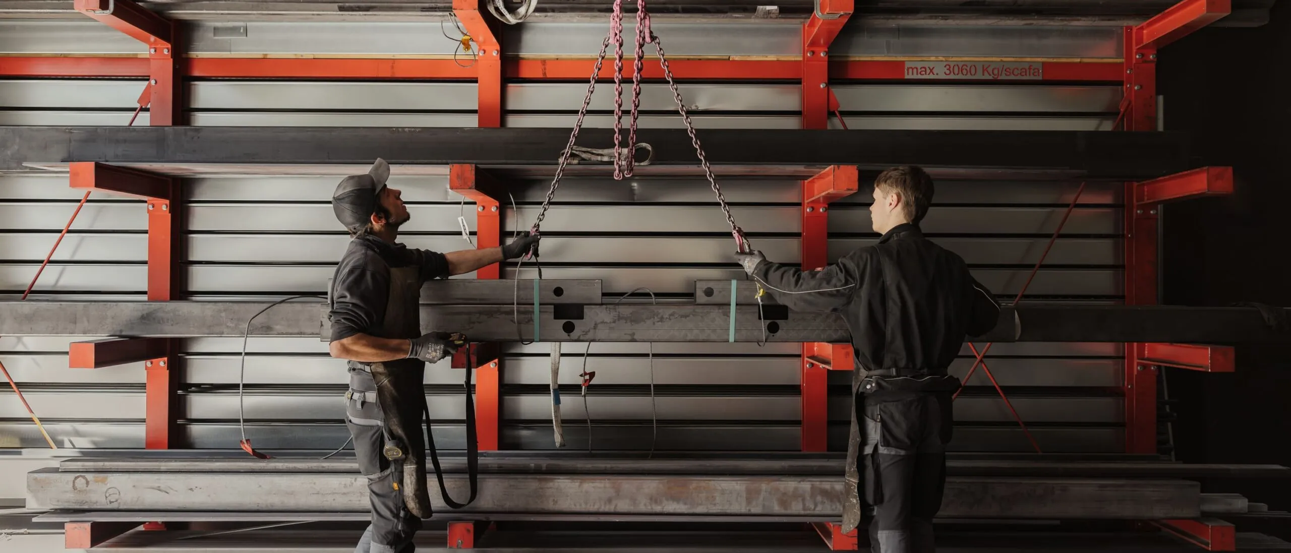 Zwei Mitarbeitende in einer Metallbau-Werkstatt heben einen langen Stahlträger mit einer Kette, vor roten Stahlregalen und weiteren Stahlplatten.