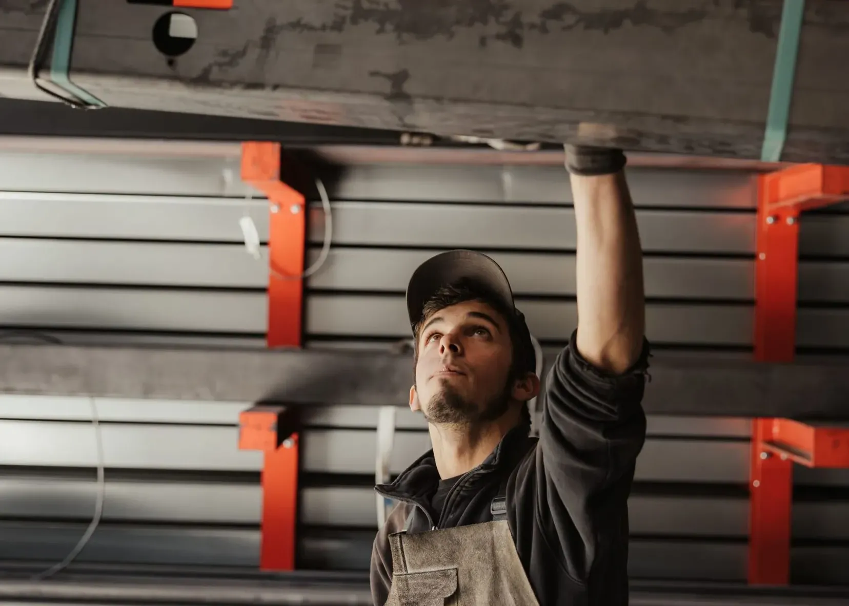 Person in Arbeitskleidung und Kappe hebt schweren Metallträger mit Kranhaken in einer Metallwerkstatt; im Hintergrund Stahlregale.