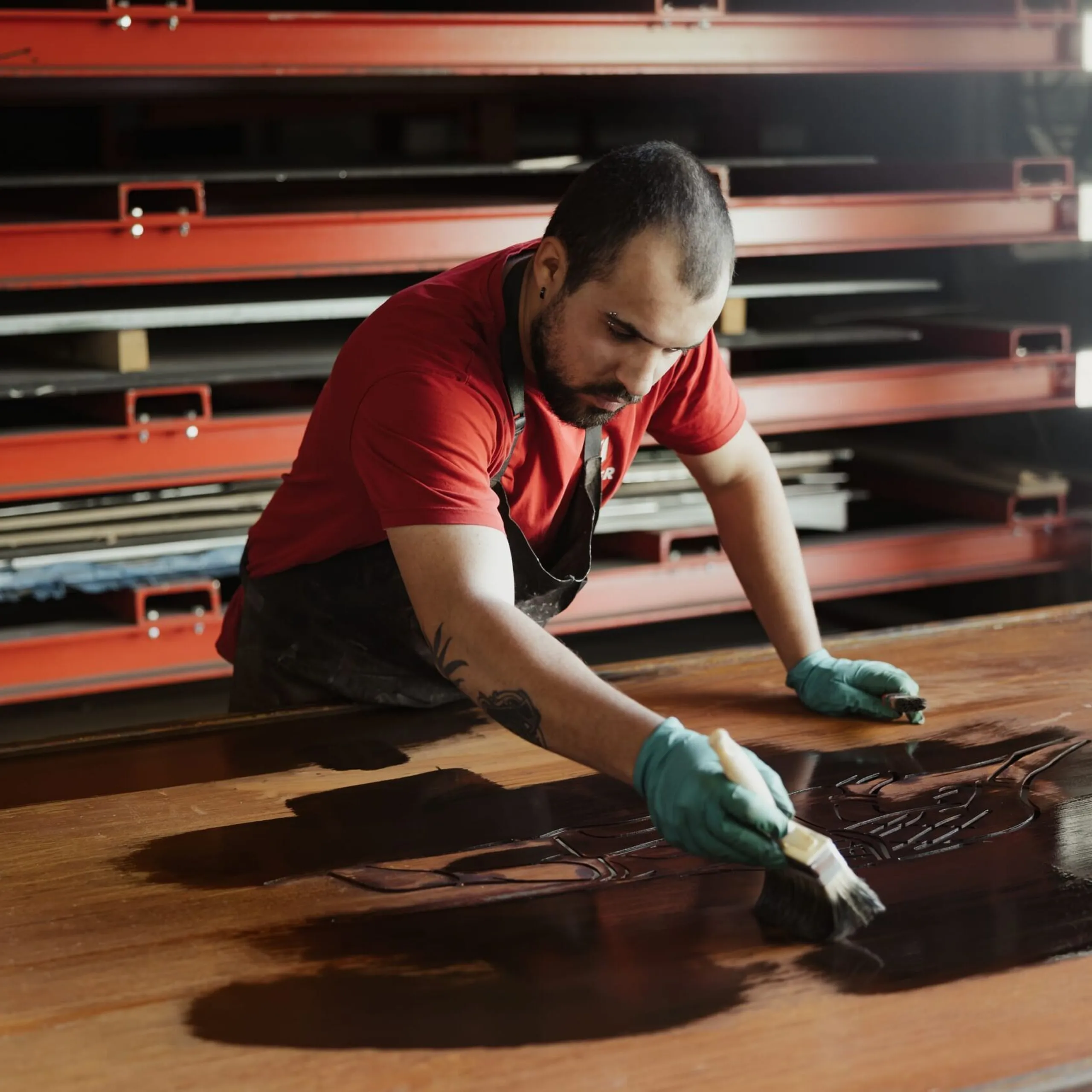 Person in rotem Shirt und grünen Handschuhen lackiert eine große Platte in einer Werkstatt; im Hintergrund rote Metallregale mit weiteren Platten.
