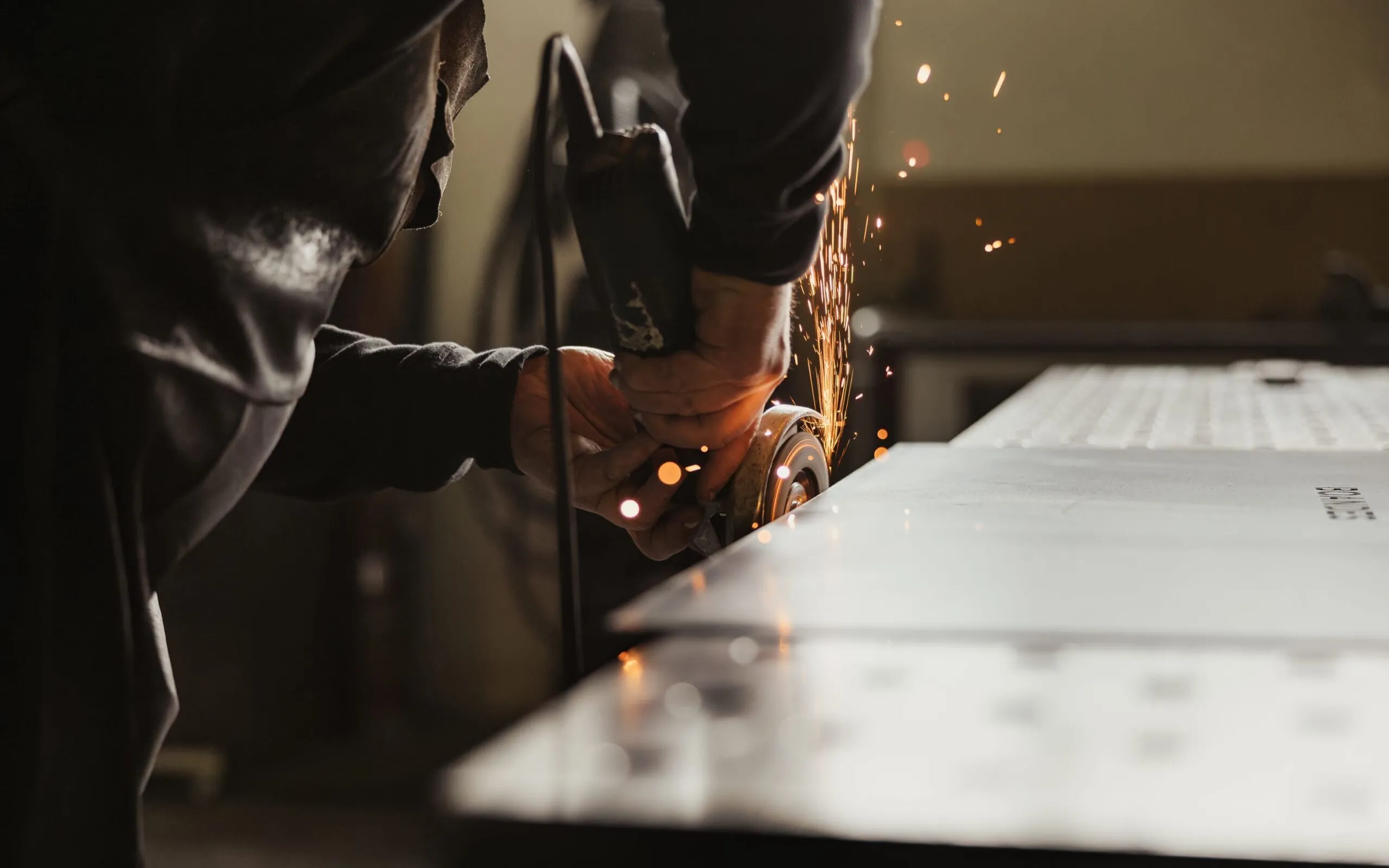 Nahaufnahme: Eine Person bearbeitet Metall mit einem Winkelschleifer; Funken sprühen, Metallblech liegt auf einer Werkbank.