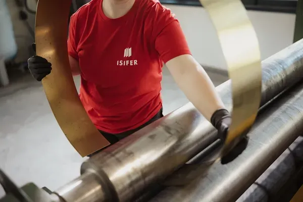 Person in rotem ISIFER-Shirt bedient eine Metallwalze und zieht ein Metallband durch eine Maschine in einer Werkstatt.