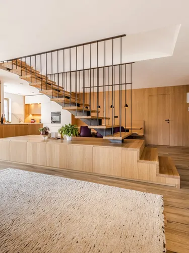 Offener Wohnbereich mit moderner Treppe: Holzstufen schweben an schwarzen Metallstangen, dahinter Küche und Holzverkleidung; Teppich und Holzboden.
