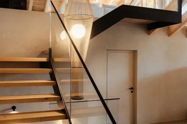 Moderne Innenansicht eines Treppenhauses: Holzstufen, Glasgeländer, Metallkonstruktion und große Pendelleuchte, Holzbalkendecke, offene Galerie.