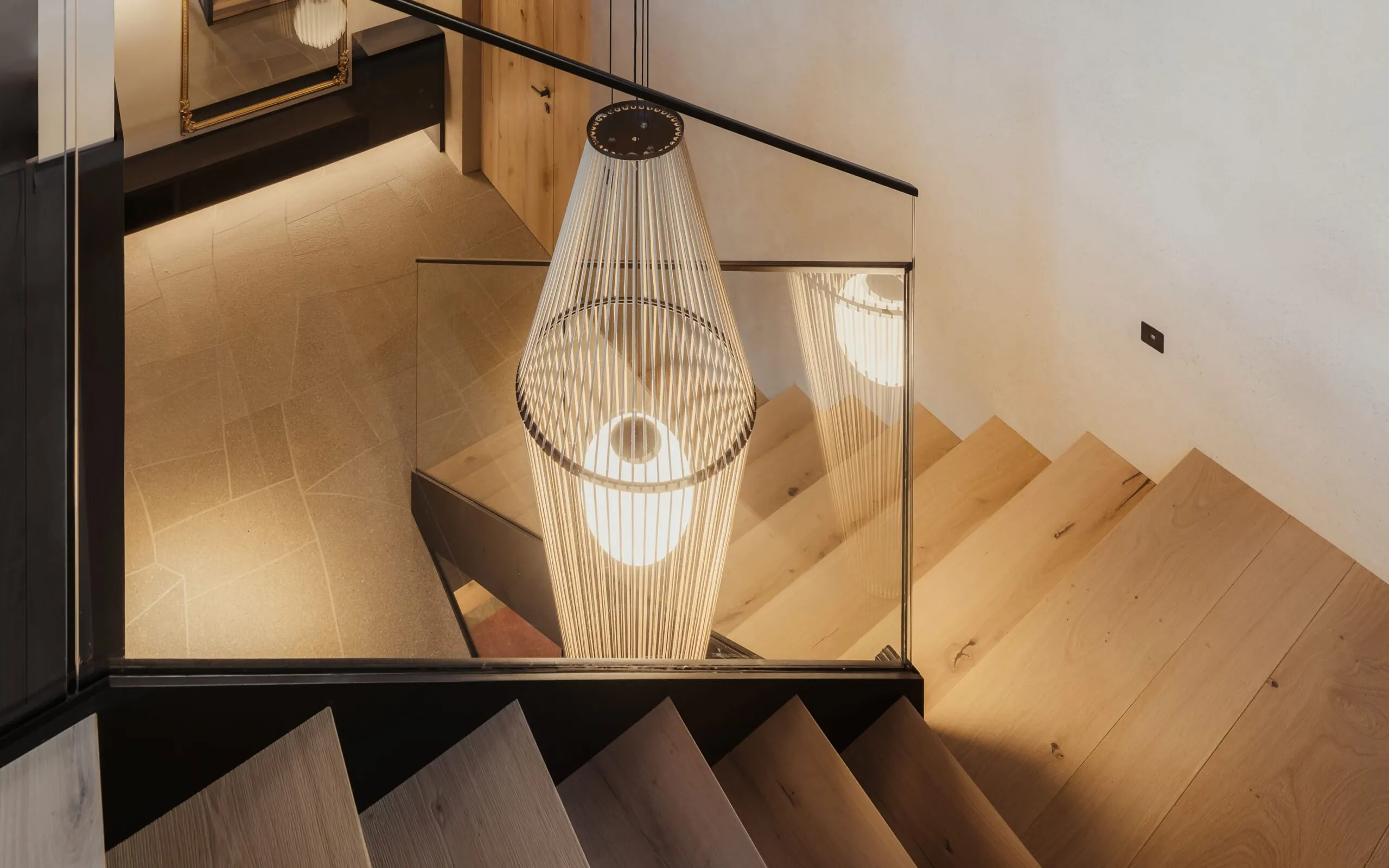 Moderne Holztreppe mit Glasgeländer; von oben Blick auf eine auffällige Pendellampe aus dünnen Metallfäden, warm beleuchtet.