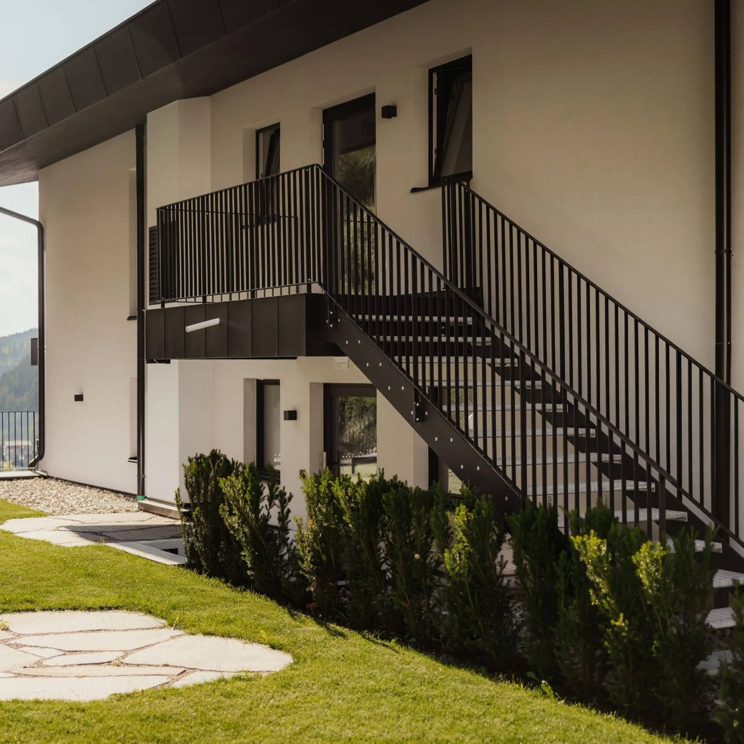 Außenansicht eines modernen weißen Gebäudes mit schwarzer Metallaußentreppe zum Balkon, Geländer, gepflegtem Rasen, Steinpfad und Hecken; Bergblick im Hintergrund, Südtirol.