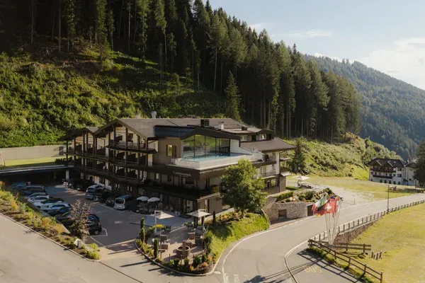 Alpenhotel aus Holz und Stein am Berghang, Terrasse mit Pool, Balkone. Parkplatz mit Autos, Wald im Hintergrund, kurvige Straße mit Schweizer Flaggen.