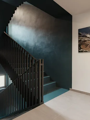 Innenansicht eines Treppenhauses mit schwarzer Metallbrüstung, dunkelblauer Wand, hellen Bodenfliesen und rechts ein Alpenlandschafts-Plakat.