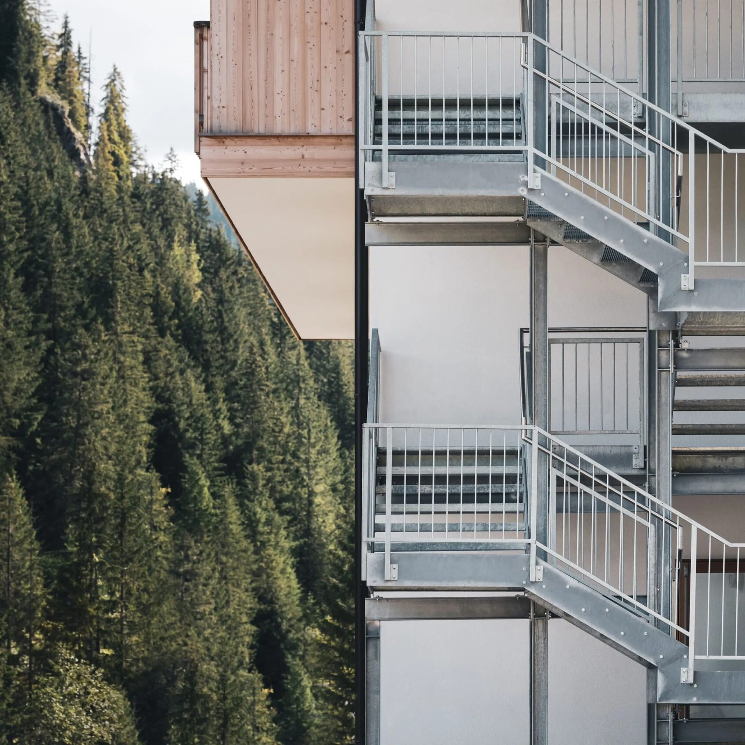 Außenmetalltreppe an einem weißen, mehrstöckigen Gebäudeteil; Stahlgeländer, linker Hintergrund dichter Nadelwald.