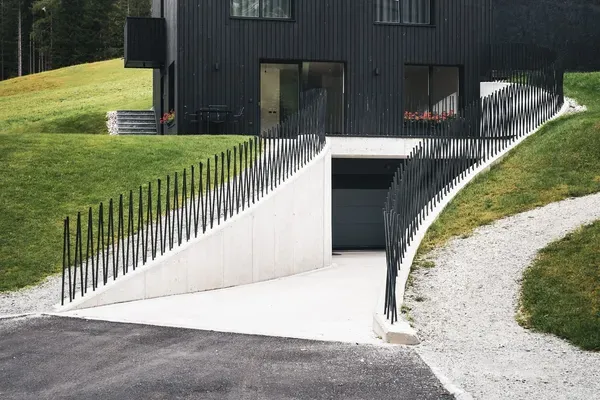 Modernes, schwarzes Metallhaus auf Hanglage in Südtirol; große Fensterfronten, Betonrampe mit spitzen Metallzäunen, grüne Wiesen und Wald im Hintergrund.