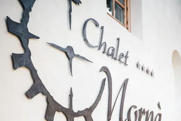 Außenansicht einer weißen Gebäudewand mit Metallschriftzug Chalet Morna; daneben eine alte Holzleiter und ein Fenster mit rotem Blumenkasten.