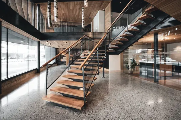 Moderne Lobby einer Südtiroler Metallbaufirma: offene Holzstufen-Treppe mit Metallrahmen und Netzgeländer, Glaswände, Pflanzen und warmes Licht.