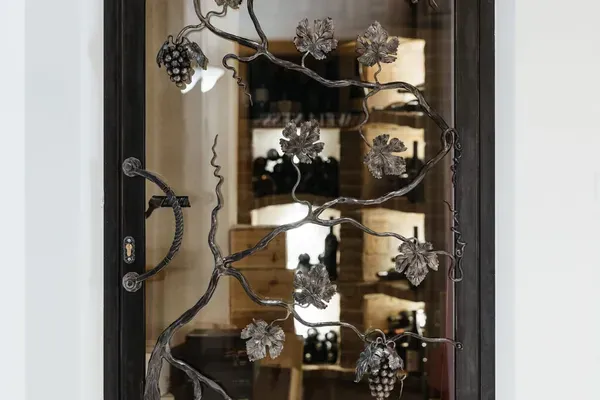 Metalltür mit dekorativem Traubenranken-Gitter, Blätter und Trauben; Glasfenster dahinter zeigt Weinregale; unten das Wort VIN.