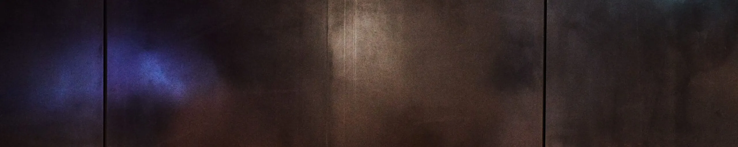 Metallische Wand aus großen Paneelen mit senkrechten Fugen; reflektierende Oberfläche mit blauen, türkisen und warm orangefarbenen Lichtreflexen.