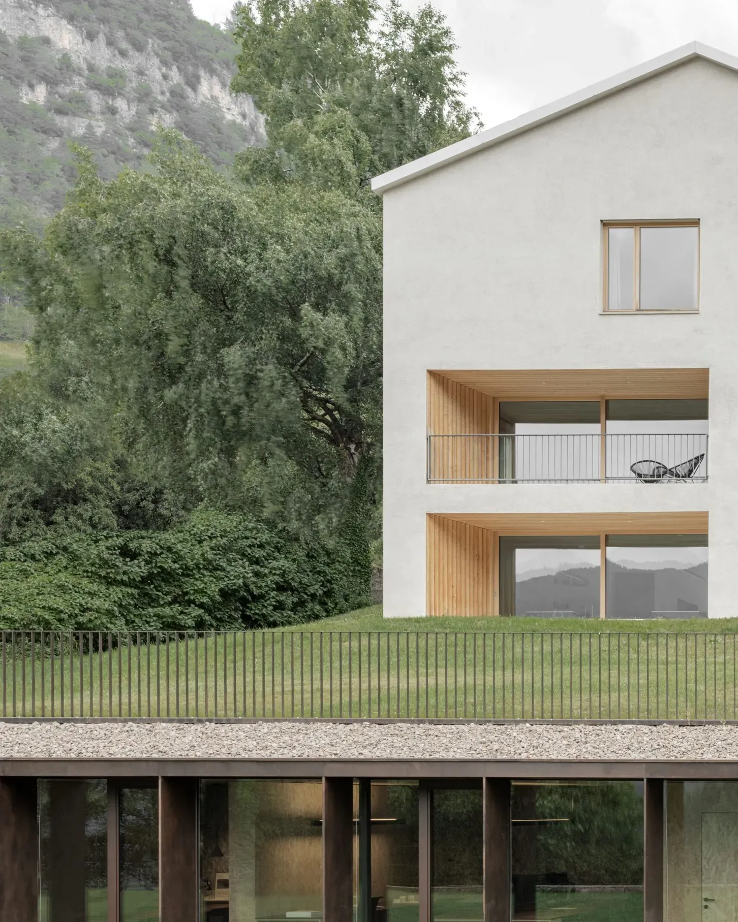 Casa moderna con facciata bianca, terrazze in legno e parapetti in metallo, vetrate al piano terra, circondata da verde e montagne sullo sfondo.