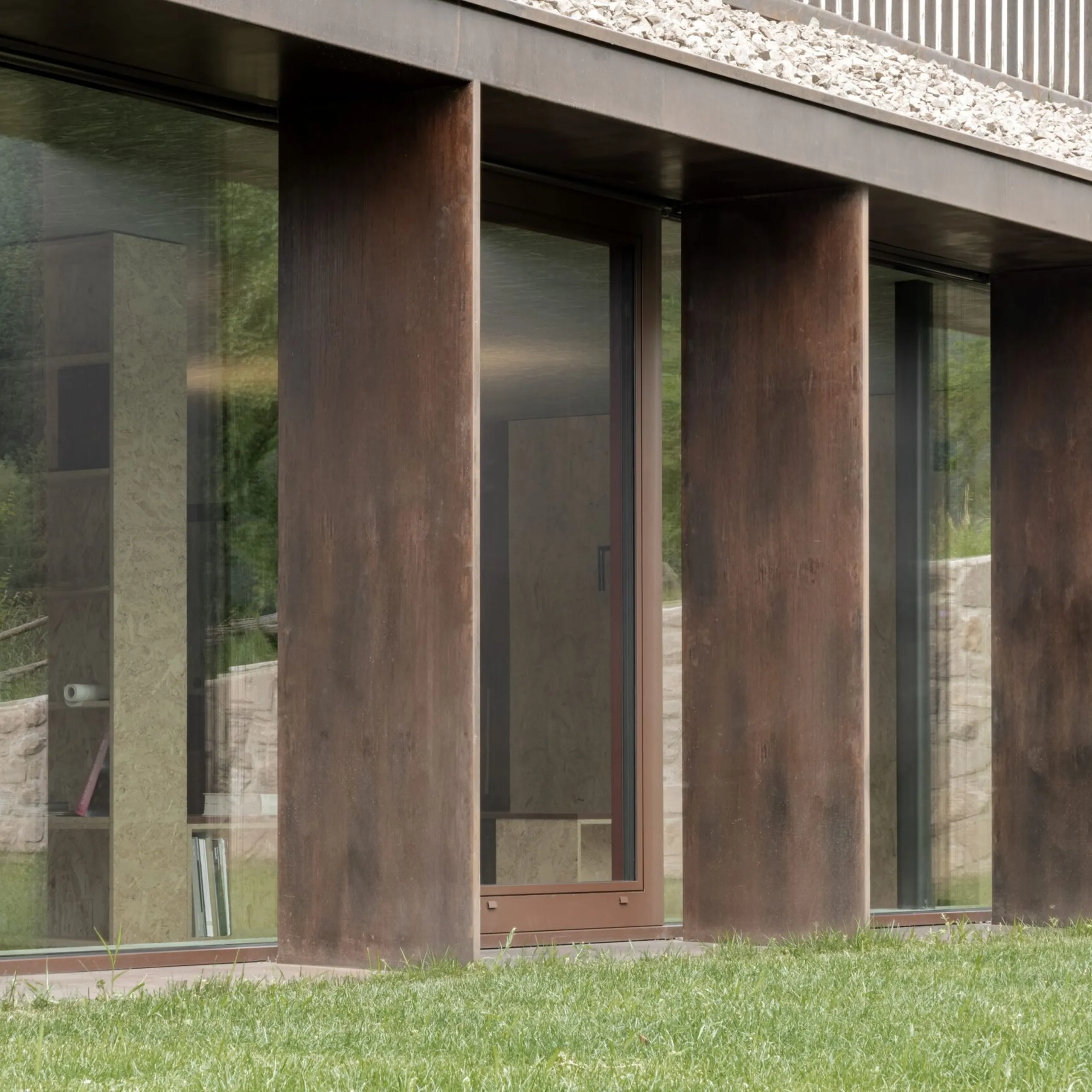 Facciata moderna di un edificio industriale: colonne in acciaio corten tra grandi vetrate, prato verde davanti e bordo in pietra.