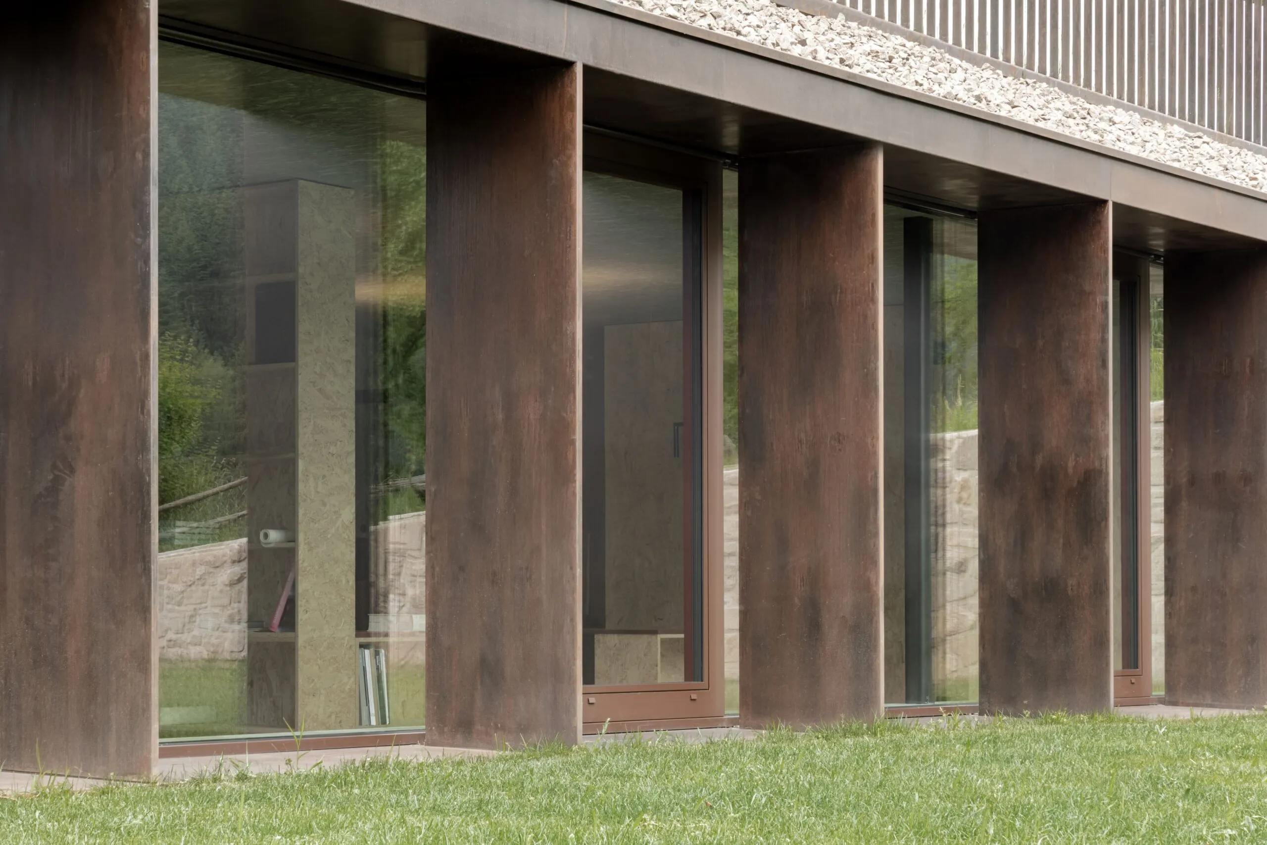 Facciata moderna di un edificio industriale: colonne in acciaio corten tra grandi vetrate, prato verde davanti e bordo in pietra.