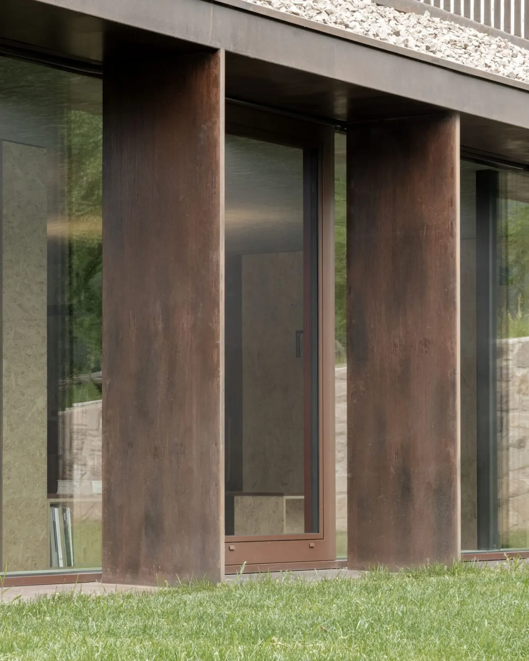 Facciata moderna di un edificio industriale: colonne in acciaio corten tra grandi vetrate, prato verde davanti e bordo in pietra.