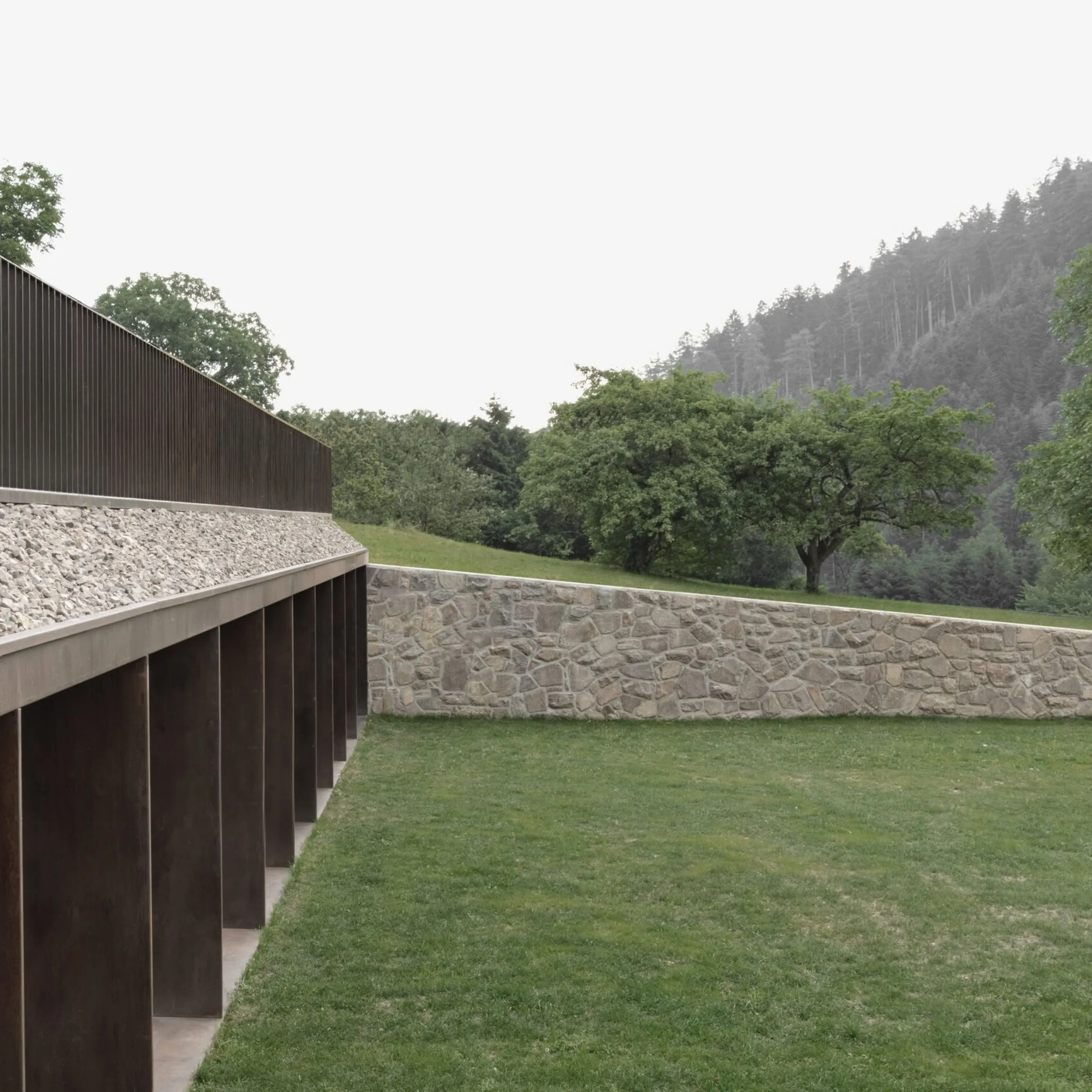 Lunga barriera metallica con barre verticali su una sporgenza di ghiaia, accanto a una parete di pietra; prato verde, alberi e foresta montuosa sullo sfondo.