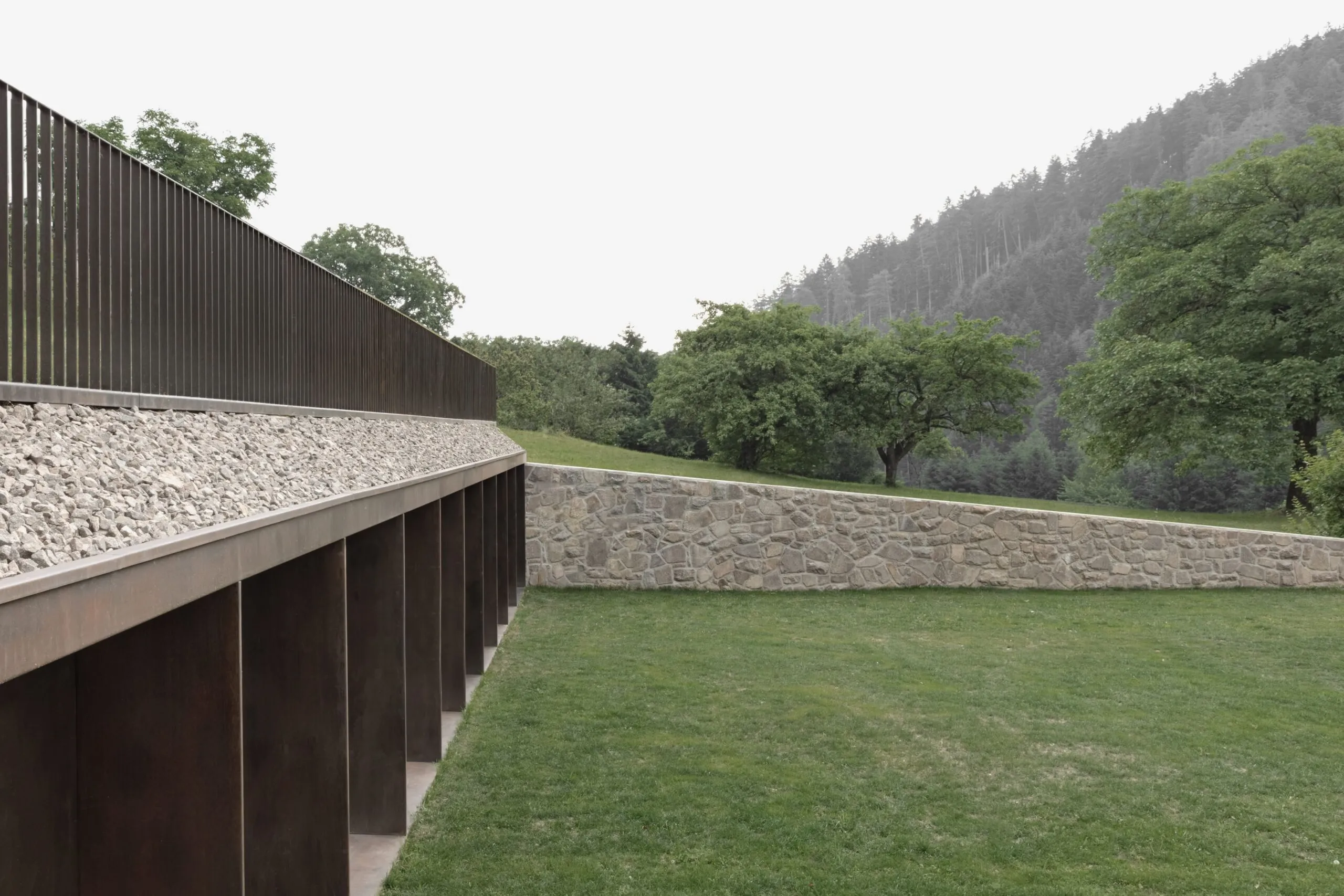 Lunga barriera metallica con barre verticali su una sporgenza di ghiaia, accanto a una parete di pietra; prato verde, alberi e foresta montuosa sullo sfondo.