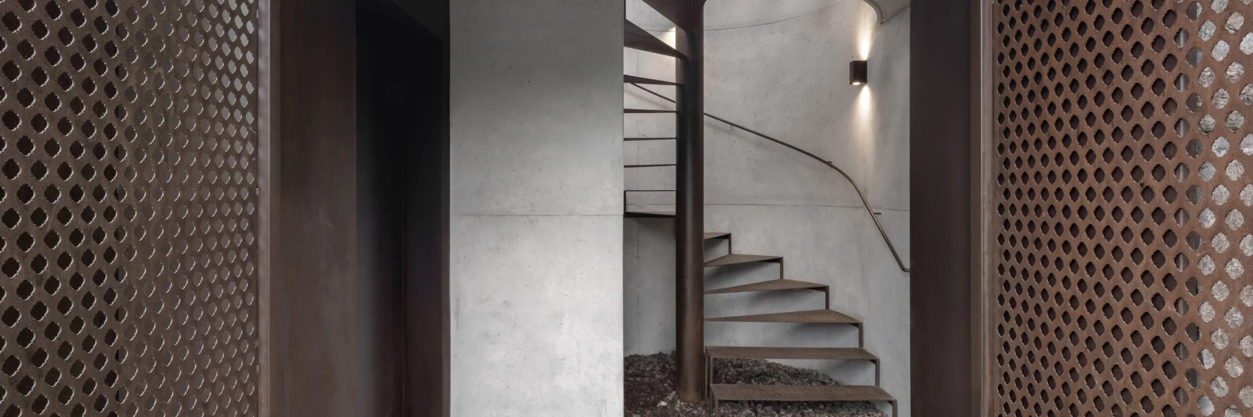 Ingresso moderno in cemento e metallo: scala a chiocciola in metallo attorno a una colonna, pareti chiare, pannelli laterali perforati e pavimento ghiaioso.