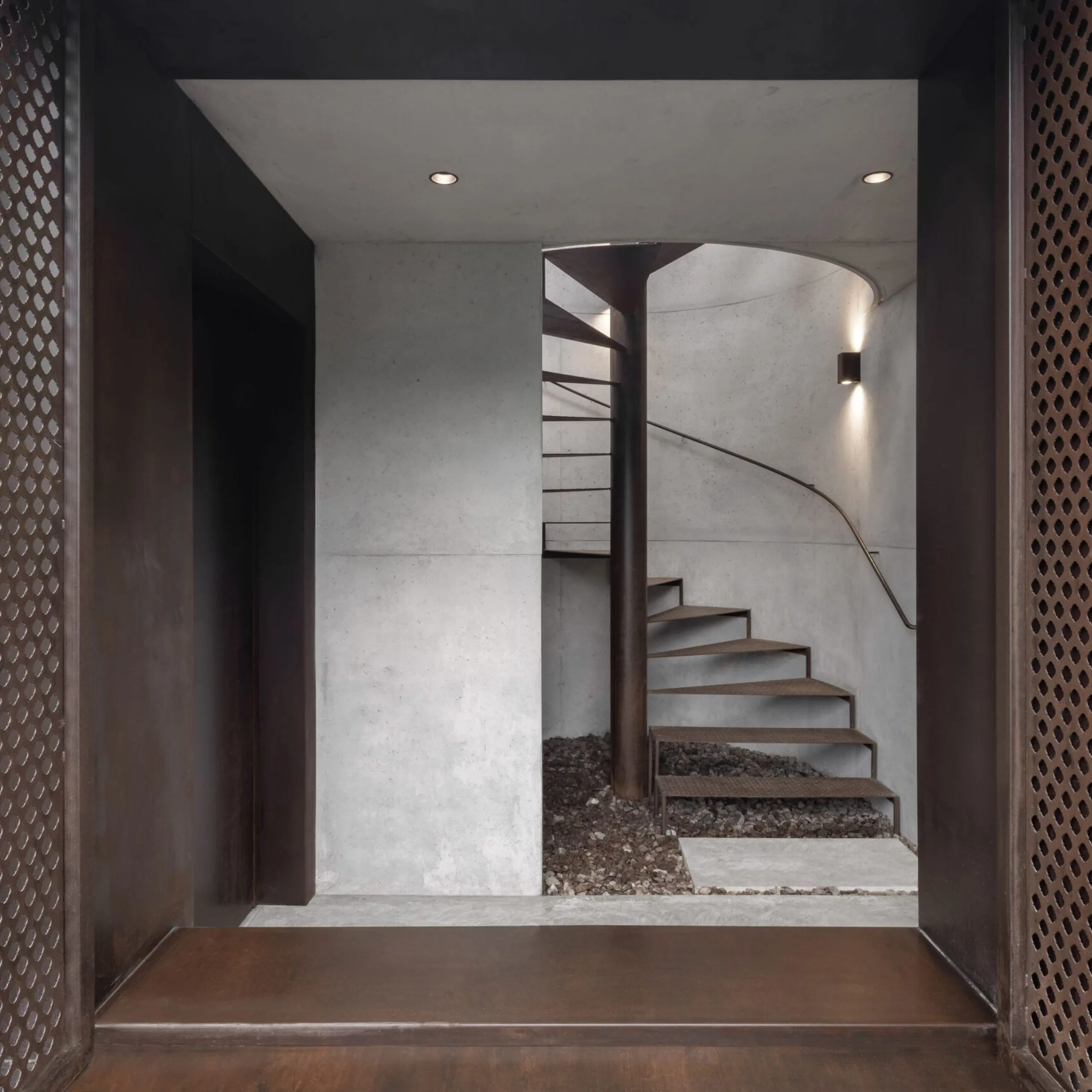Ingresso moderno in cemento e metallo: scala a chiocciola in metallo attorno a una colonna, pareti chiare, pannelli laterali perforati e pavimento ghiaioso.