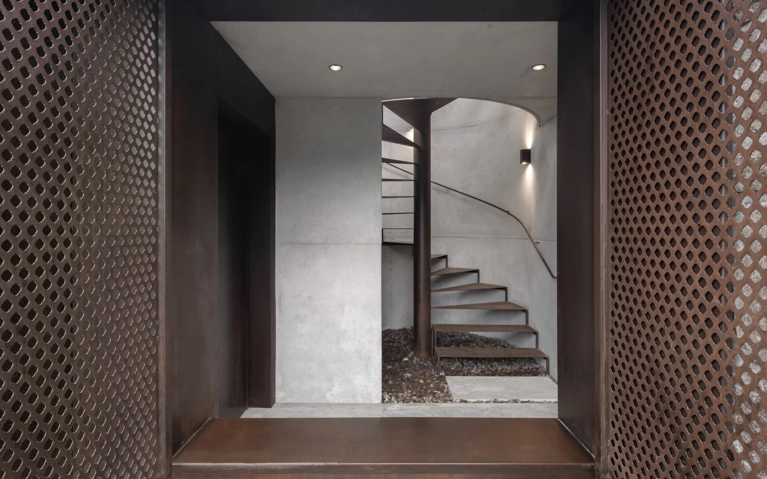 Ingresso moderno in cemento e metallo: scala a chiocciola in metallo attorno a una colonna, pareti chiare, pannelli laterali perforati e pavimento ghiaioso.