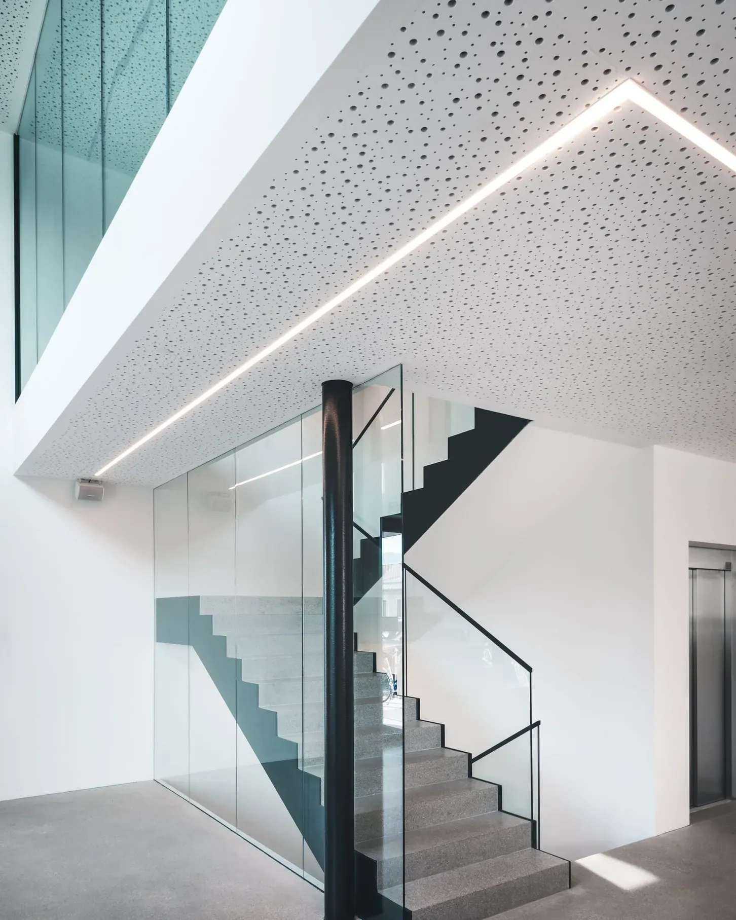 Moderne Innenansicht eines Glas- und Metallbaus: Treppe mit Glasgeländer, schwarzer Stützpfosten, transparente Glaswände und perforierte Decke mit LED-Lichtlinie.
