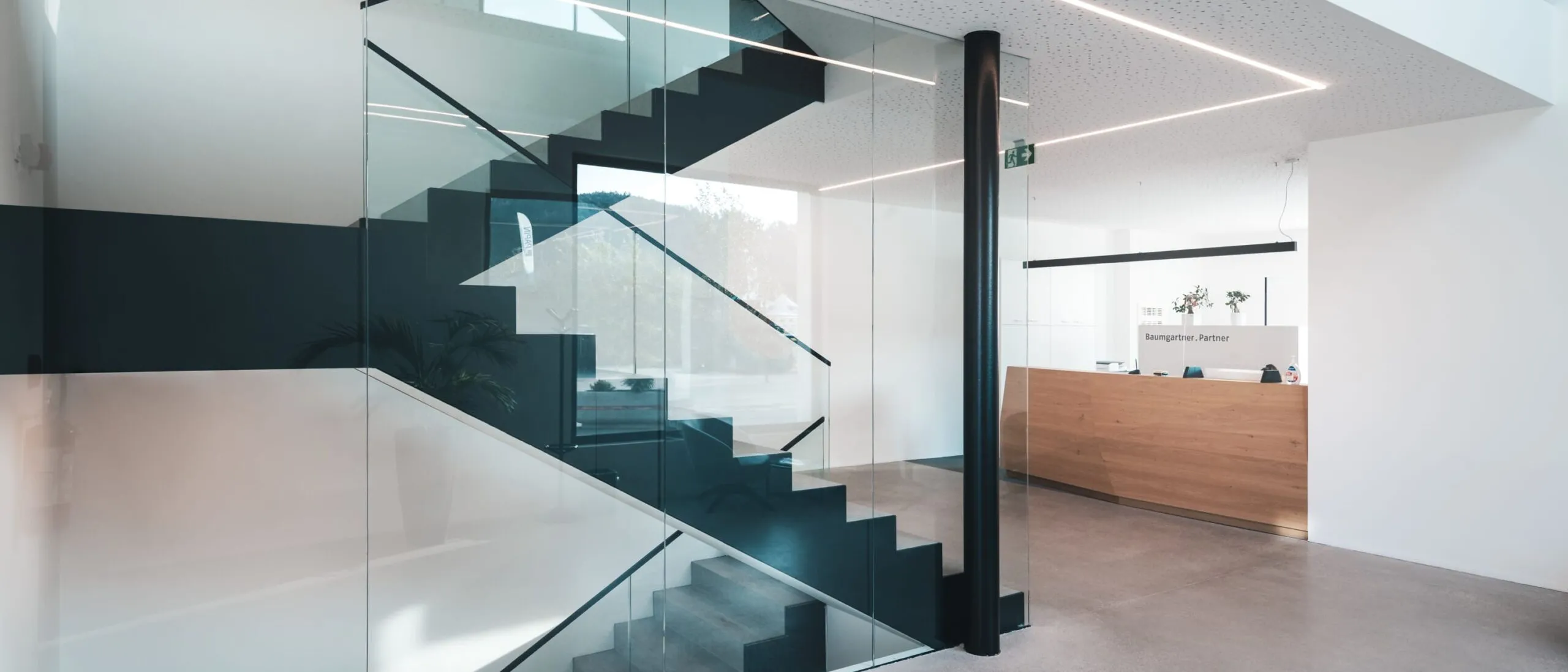 Moderne Lobby einer Metallbau-Firma in Südtirol: Glasfront, offene Treppe mit dunklen Stufen, heller Innenraum, Holz-Rezeption im Hintergrund.