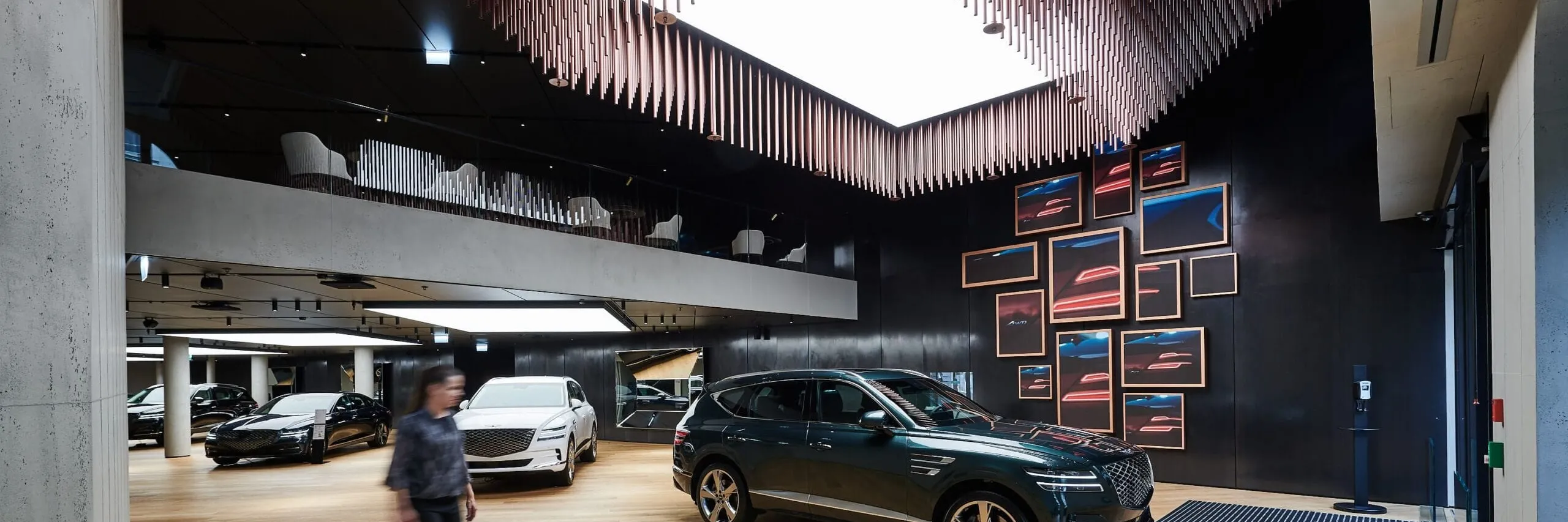 Interno showroom auto altoatesino: pavimento in legno, SUV scuro in primo piano, altre auto sullo sfondo, parete nera con quadri e lampadario a listelli.