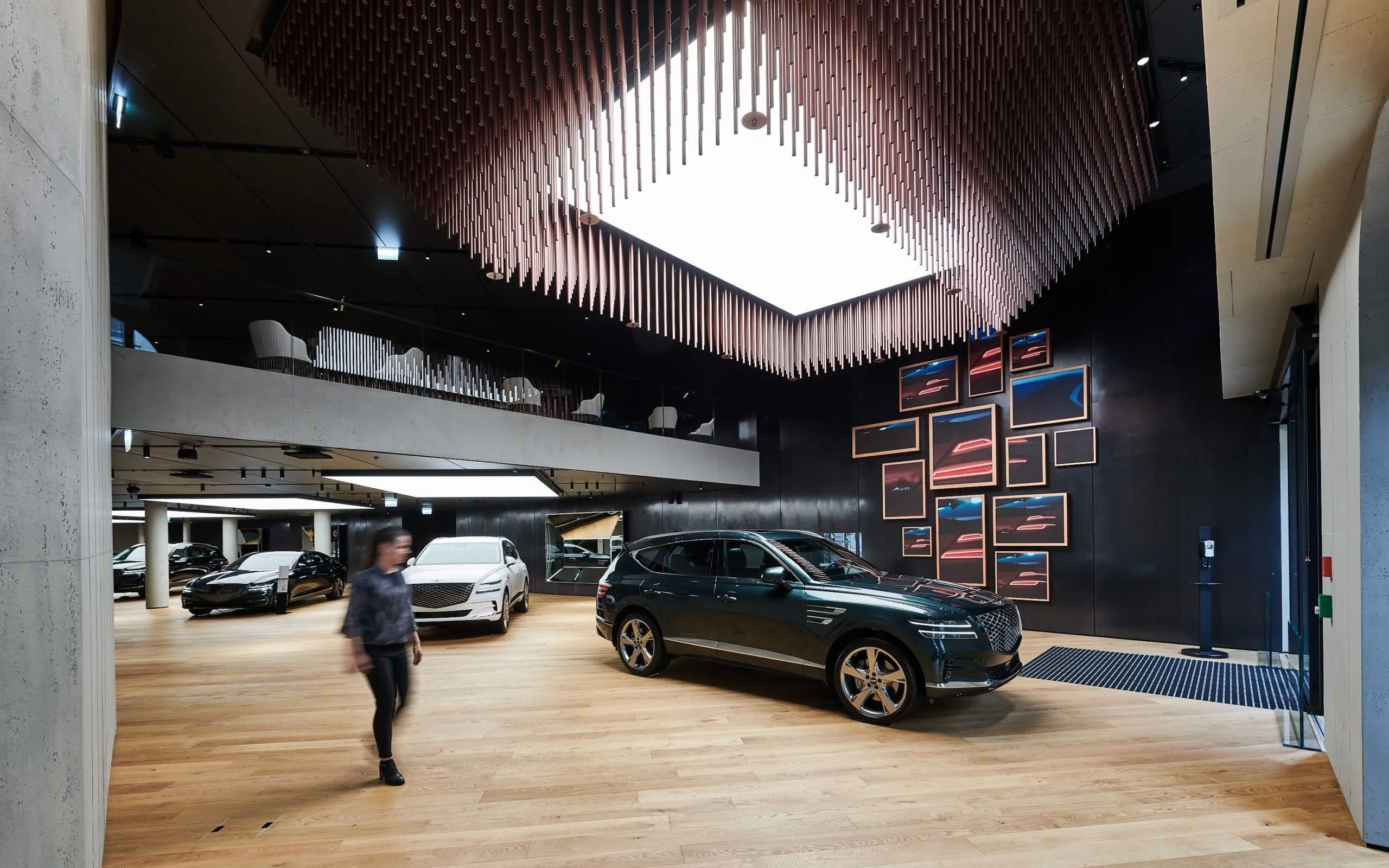 Showroom eines Metallbau-Unternehmens in Südtirol: Autos auf Holzboden, dunkle Wand mit Bilderrahmen, auffällige Deckenlamellen; eine Person geht vorbei.