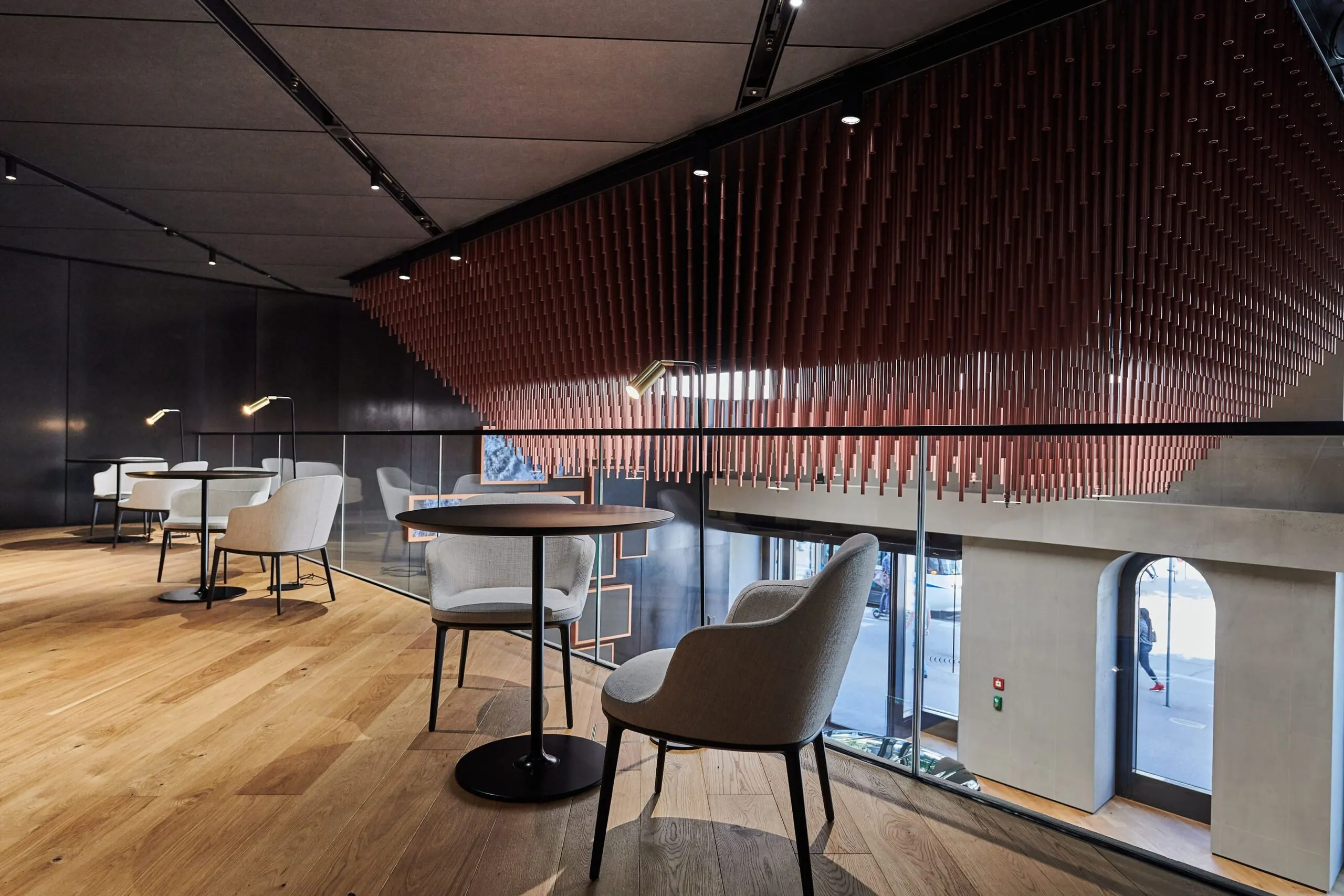 Loungebereich eines Metallbau-Unternehmens: Holzfußboden, runde Tische mit Stühlen; kupferfarbene Skulptur aus senkrechten Streben hängt von der Decke.