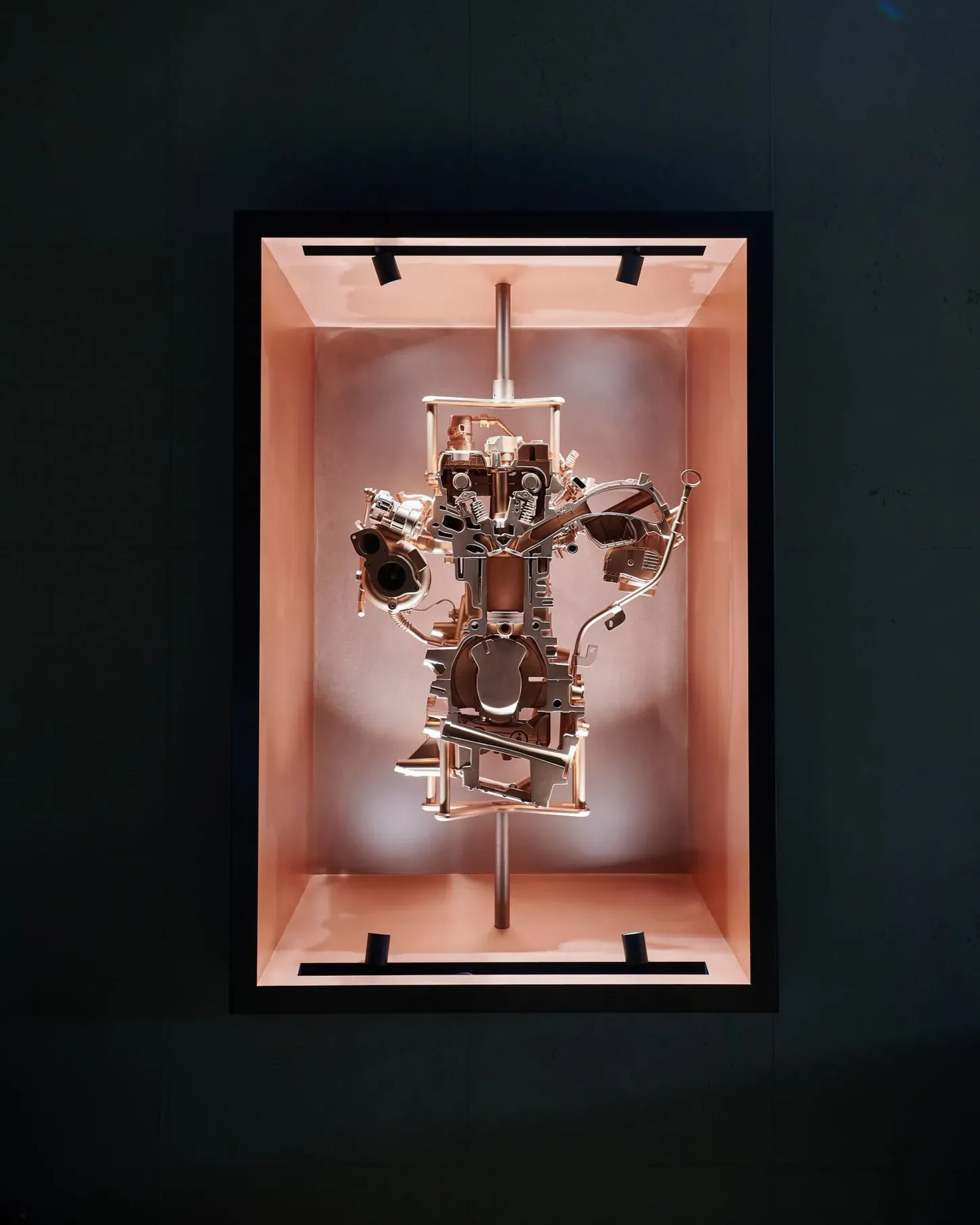 Komplexe Metall-Motorbaugruppe in einem beleuchteten kupferfarbenen Ausstellungskasten, vor dunkler Wand.