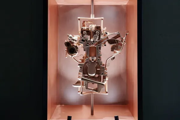 Komplexe Metall-Motorbaugruppe in einem beleuchteten kupferfarbenen Ausstellungskasten, vor dunkler Wand.