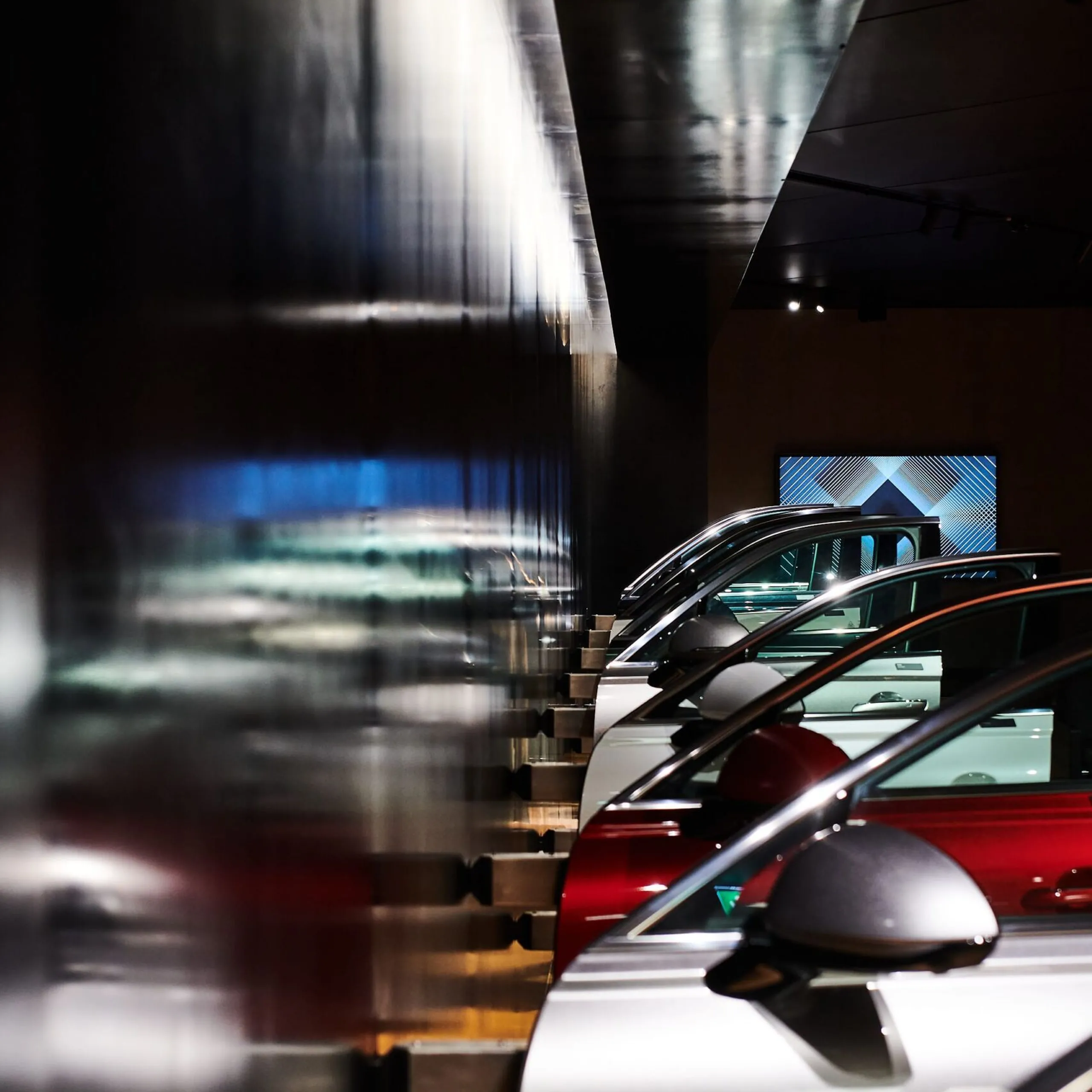 Mehrere Autos in einer dunklen Industrieausstellung, hintereinander geparkt; links reflektierende Metallwand, rechts Fahrzeugfronten sichtbar.