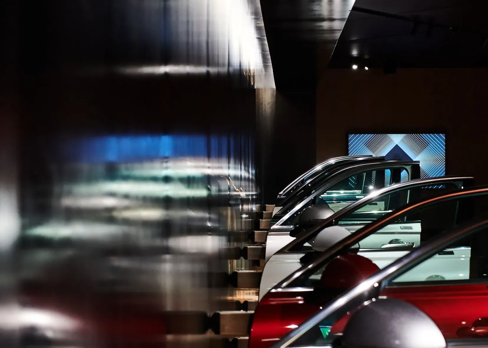 Mehrere Autos in einer dunklen Industrieausstellung, hintereinander geparkt; links reflektierende Metallwand, rechts Fahrzeugfronten sichtbar.