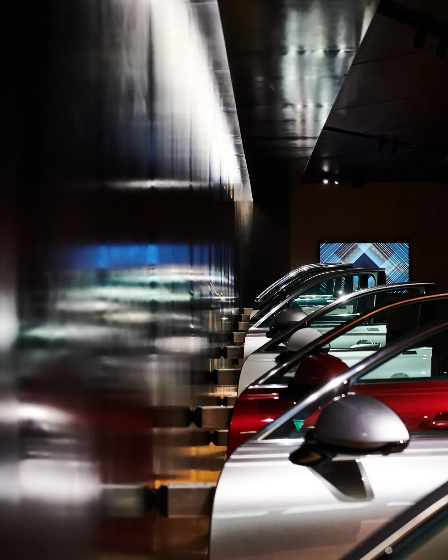 Mehrere Autos in einer dunklen Industrieausstellung, hintereinander geparkt; links reflektierende Metallwand, rechts Fahrzeugfronten sichtbar.