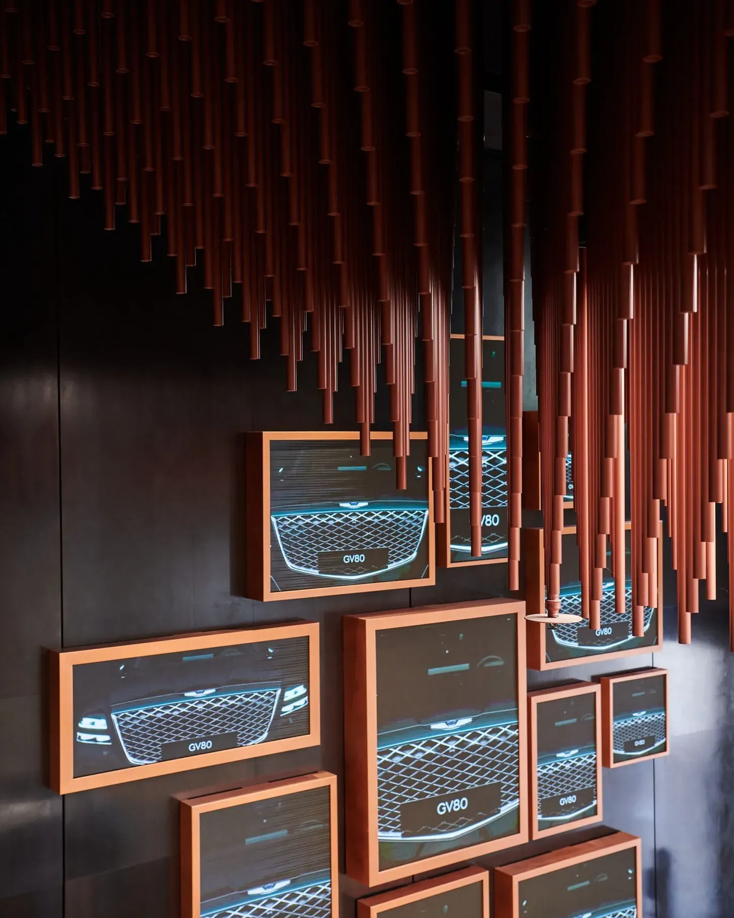 Dunkler Ausstellungsraum mit roten Metallröhren von der Decke; Holzrahmen-Bildschirme zeigen GV80-Vordergrills eines Autos.