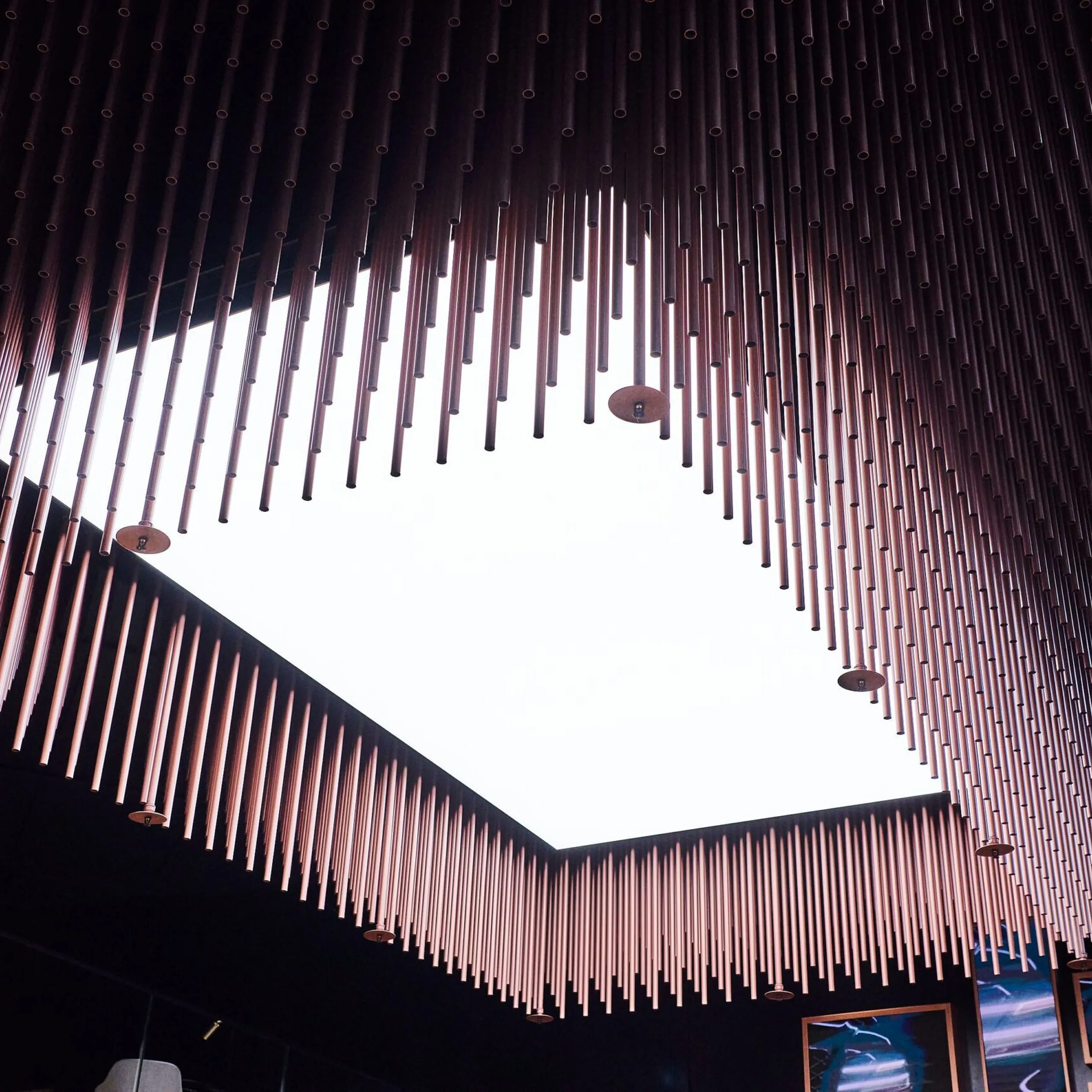 Innenraum eines Südtiroler Metallbau-Unternehmens: Dicht an der Decke hängen Metallstäbe, die eine helle quadratische Dachlichtöffnung umranden; dunkle Wände, Glasfronten.