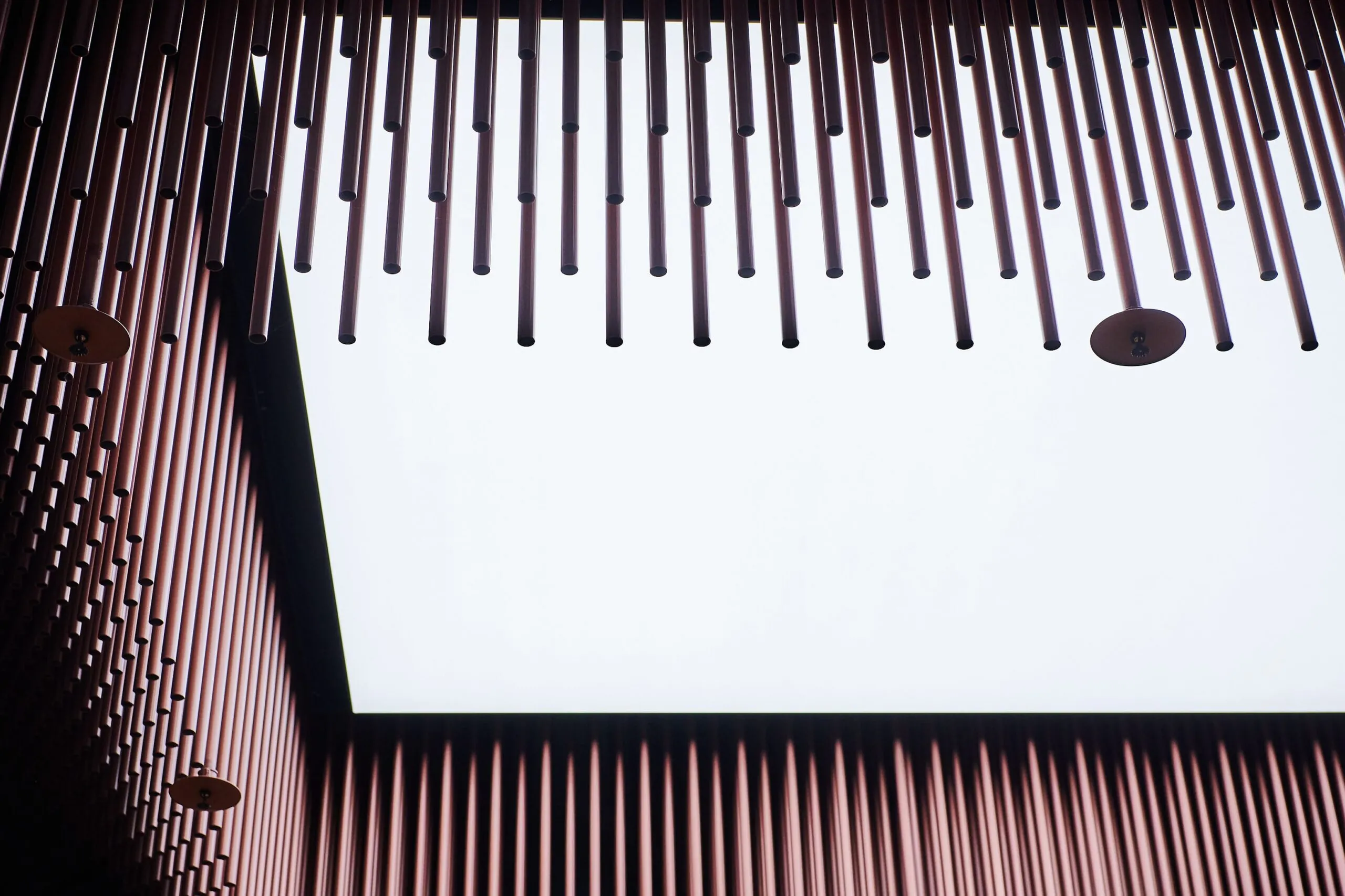 Blick von unten auf eine Deckeninstallation aus vielen kupferfarbigen Metallstangen, die eine helle quadratische Lichtöffnung einrahmen.