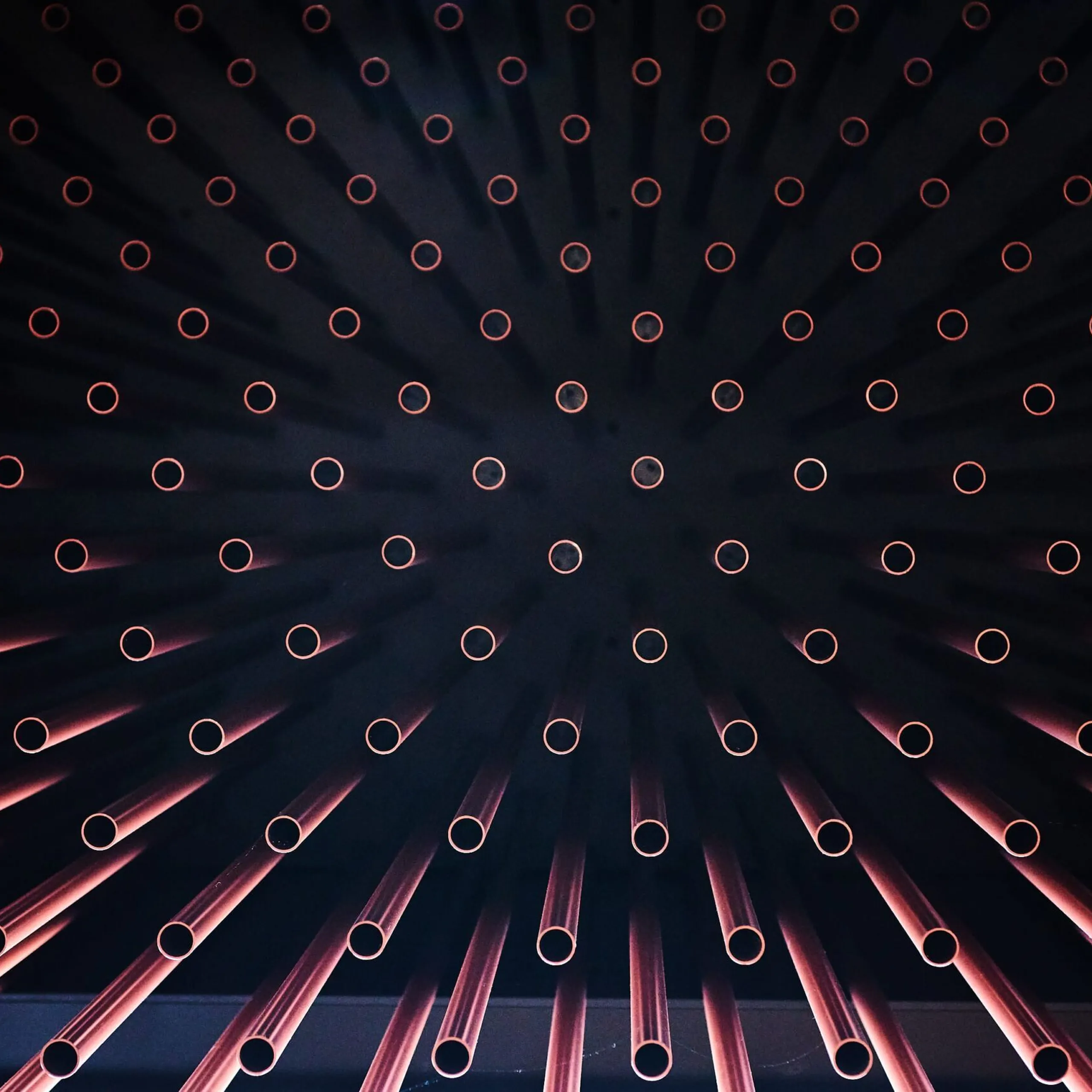 Radiale Anordnung von Metallrohren mit roten Endringen vor dunklem Hintergrund; wirkt wie Sonnenstrahlen.