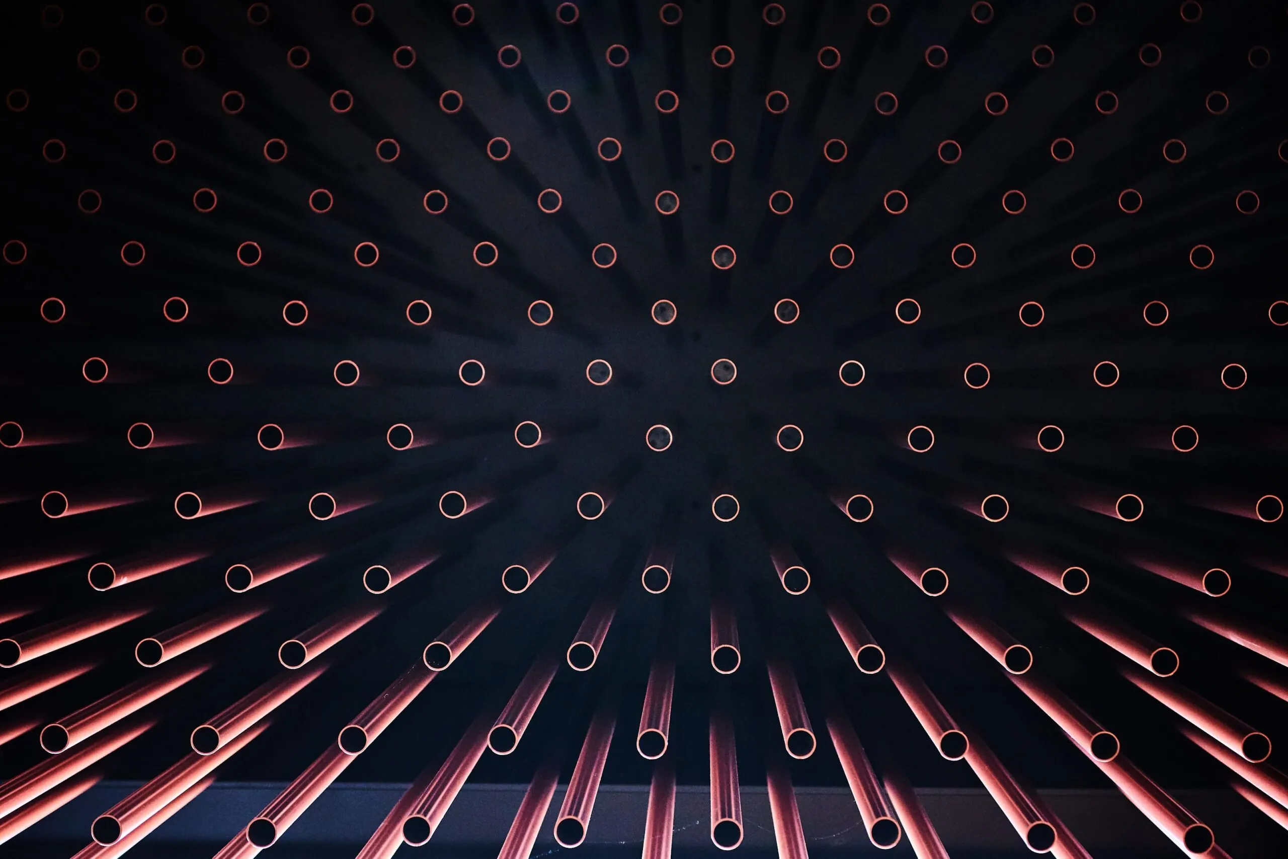 Radiale Anordnung von Metallrohren mit roten Endringen vor dunklem Hintergrund; wirkt wie Sonnenstrahlen.