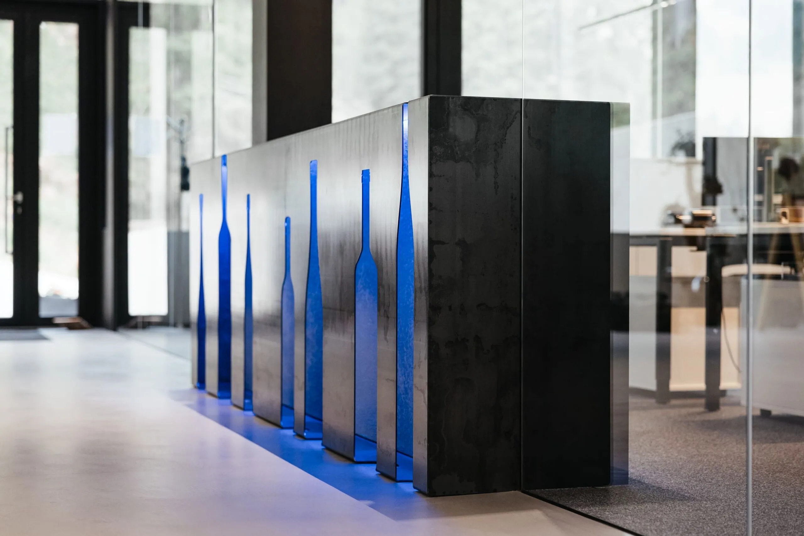 Lange schwarze Metall-Theke mit blauen Flaschenformen als Aussparungen; unten blau beleuchtet, in modernem Büro-Showroom einer Metallbau-Firma in Südtirol.