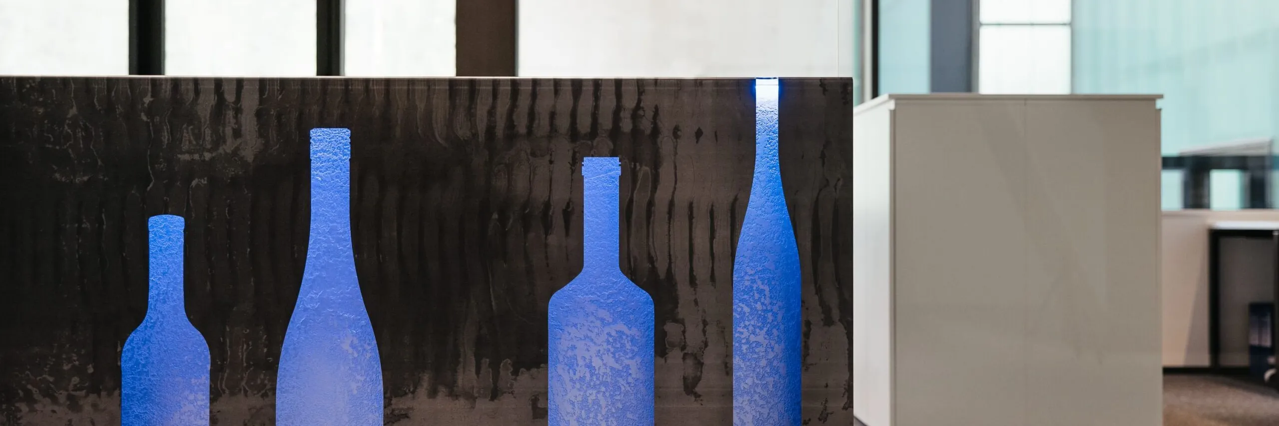 Bürobereich einer Metallbaufirma aus Südtirol: schwarzes Wandpaneel mit blauen Flaschen-Silhouetten, weiße Schränke und Glasfronten im Hintergrund.