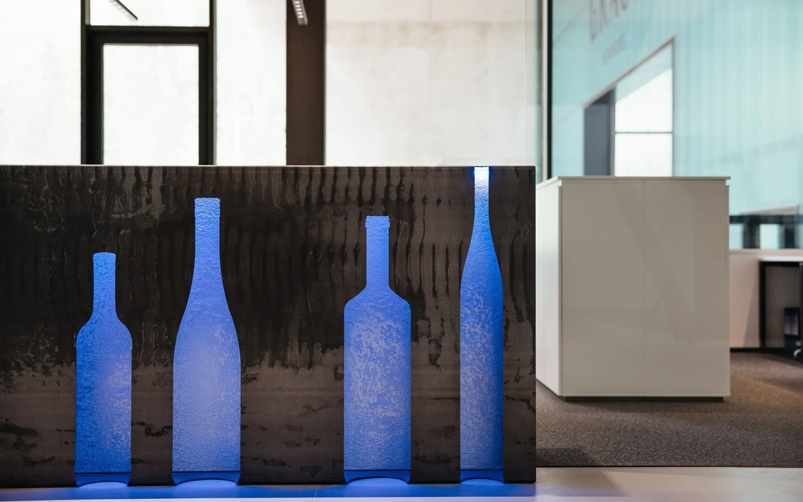 Bürobereich einer Metallbaufirma aus Südtirol: schwarzes Wandpaneel mit blauen Flaschen-Silhouetten, weiße Schränke und Glasfronten im Hintergrund.