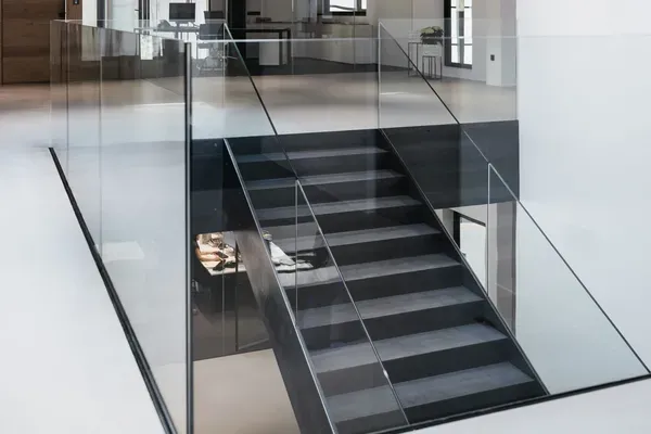 Heller moderner Bürobereich mit Glasgeländer, schwarzer Metalltreppe, weißem Boden und Oberlichtfenster; Glaswände und Arbeitsplätze im Hintergrund.